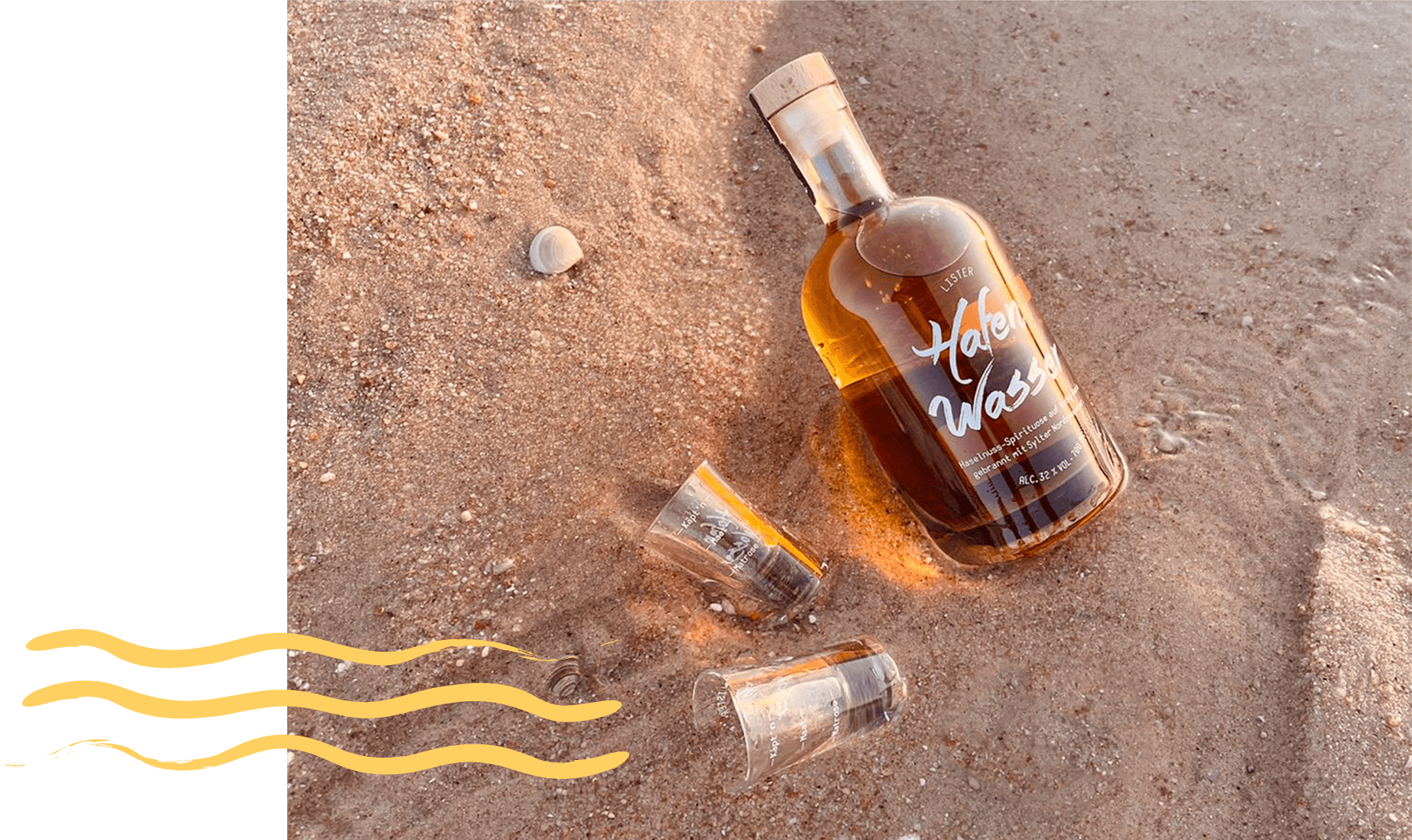Flasche Lister Hafen Wasser, Haselnuss Likör liegt im Sand auf Sylt mit zwei Shot-Gläserm